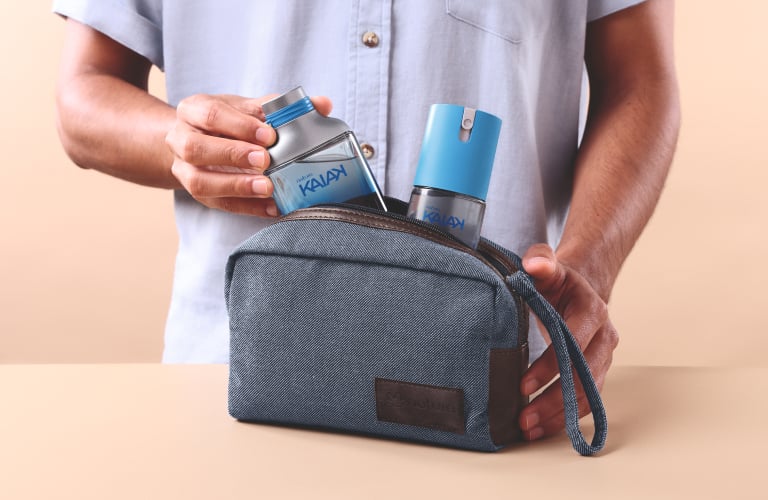 Foto com fundo bege. Mãos masculinas retirando um frasco de perfume Kaiak de uma nécessaire azul sobre uma mesa.